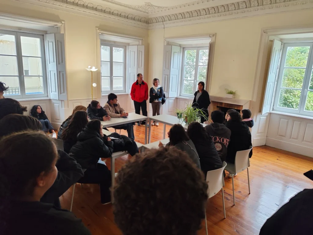 Alunos sentados em roda numa sala com janelas altas e soalho de madeira, a ouvir três adultos em pé e um sentado à mesa.