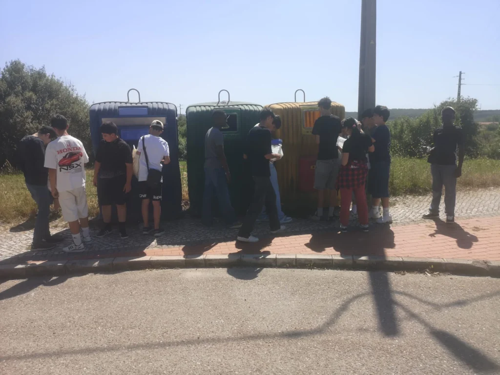 Alunos junto a três ecopontos de cores azul, verde e amarela, a depositar resíduos para reciclagem numa rua.