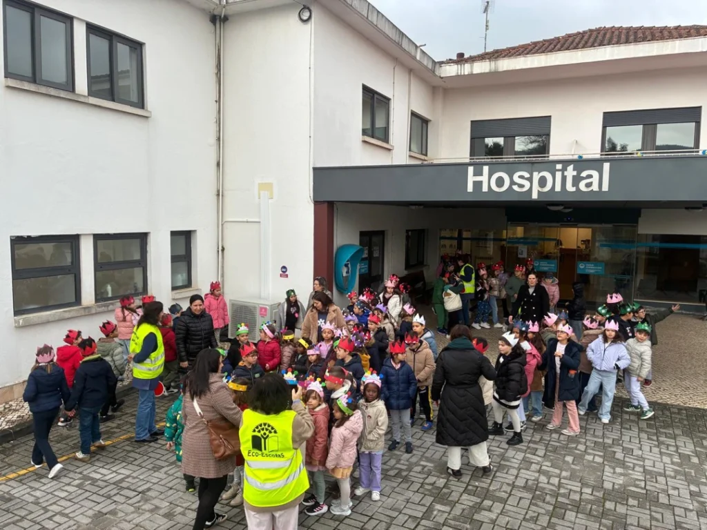 Crianças com coroas de cartão à entrada de um hospital, com um adulto de colete Eco-Escolas à frente.
