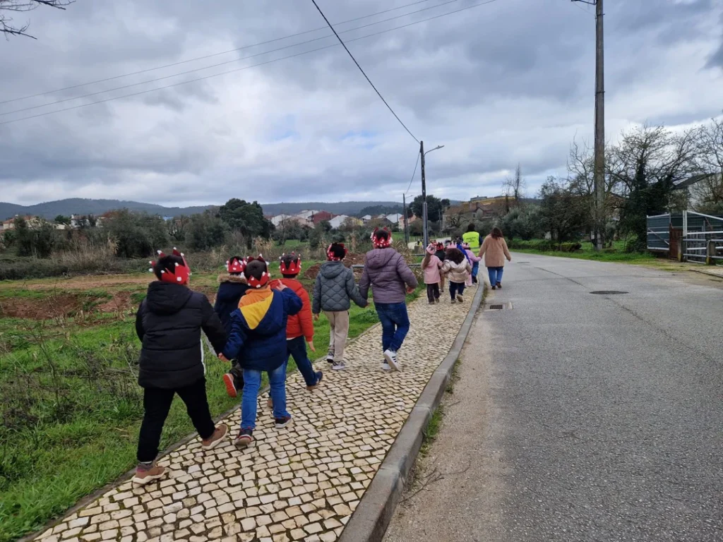 Crianças de casaco e chapéus vermelhos a caminhar em fila por um passeio de calçada junto a uma estrada.