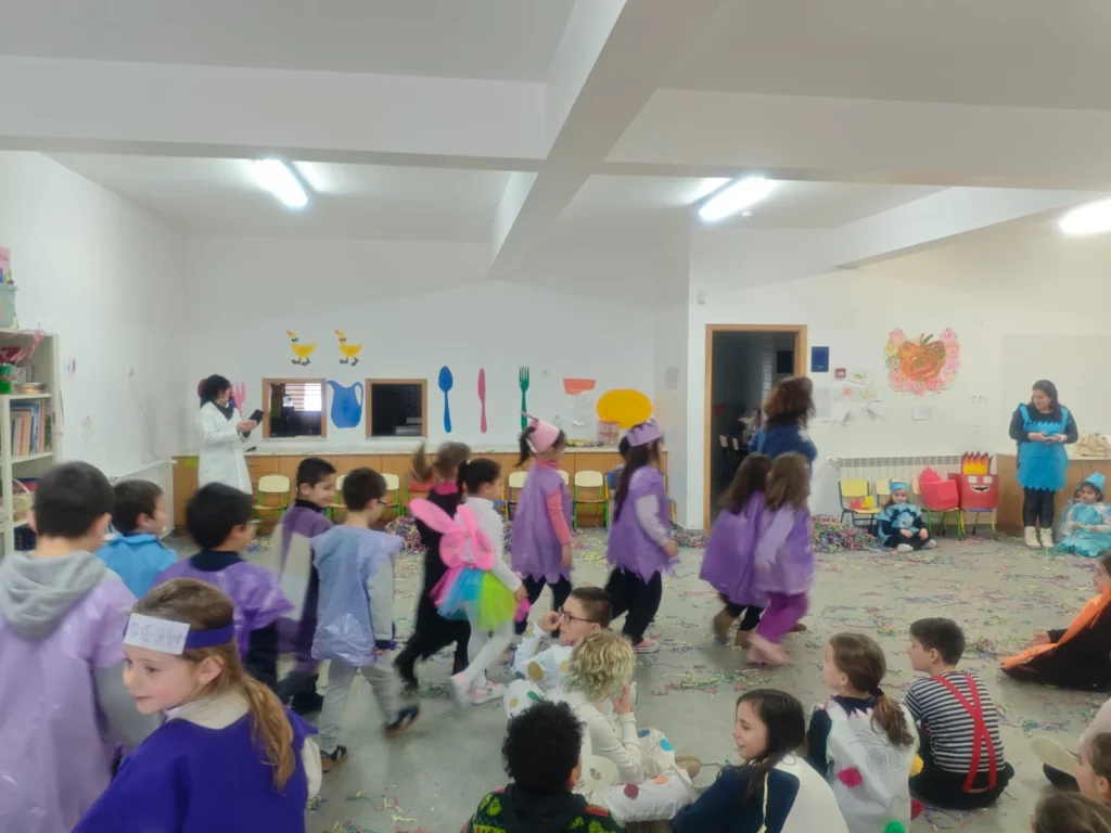 Crianças disfarçadas de Carnaval a desfilar numa sala, com outras sentadas no chão a assistir.