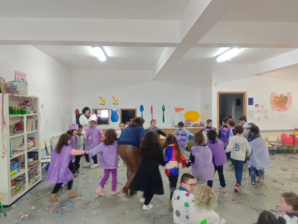 Crianças de capa lilás em roda de mãos dadas, numa sala de Carnaval com confetis no chão.