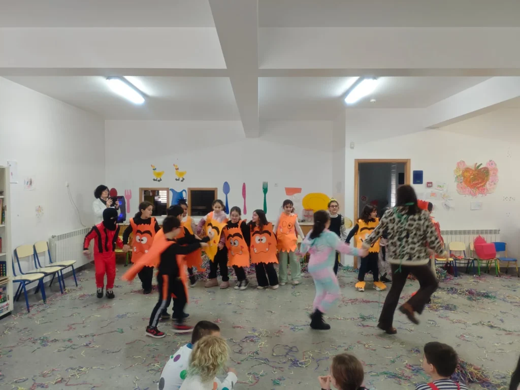 Crianças em fatos laranja de Carnaval a dançar de mãos dadas numa sala com confetis, enquanto outros assistem sentados.