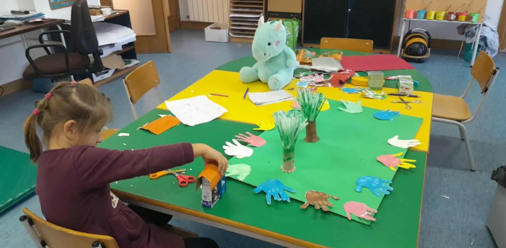 Criança a montar uma casinha de cartão numa mesa verde, com mãos e árvores de papel recortadas à volta.