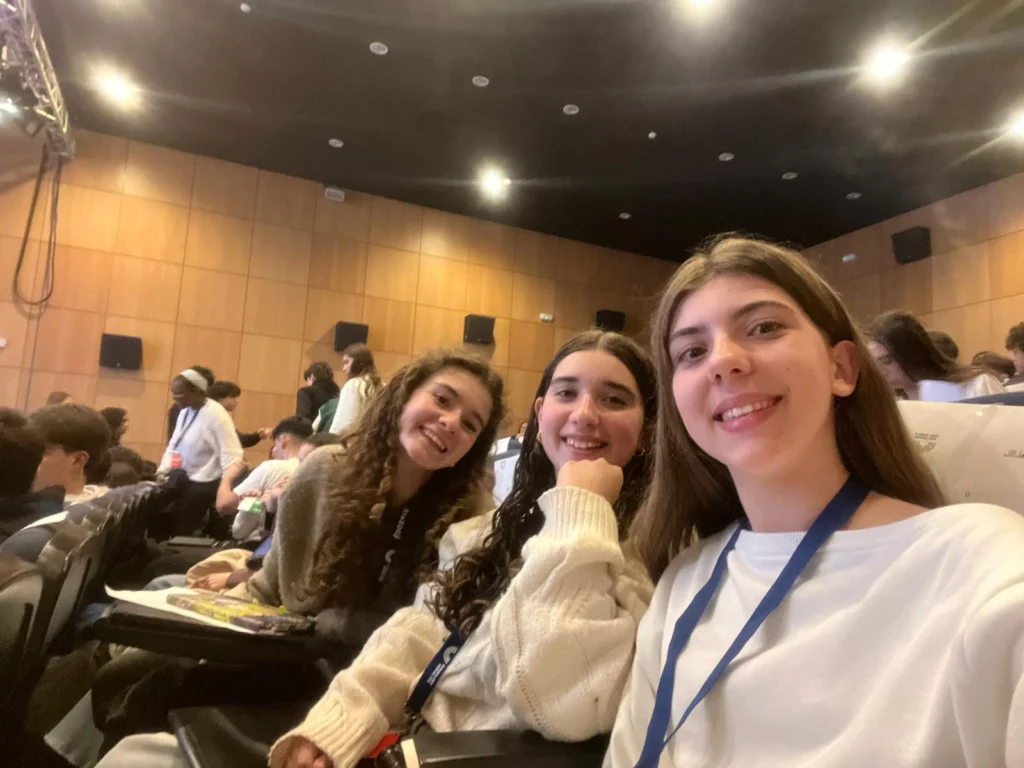 Três alunas sorriem para uma selfie, sentadas num auditório, durante a Sessão Distrital do Parlamento dos Jovens.