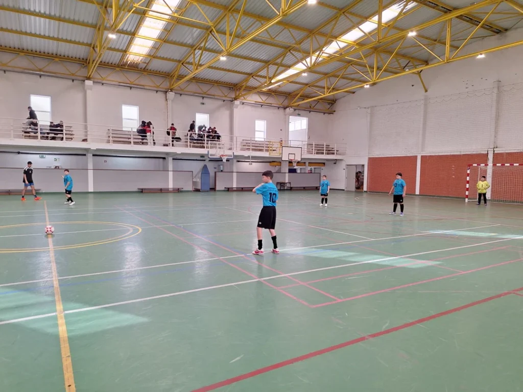 Jogadores de futsal de camisola azul espalhados por um pavilhão grande, com uma bola parada no meio-campo.