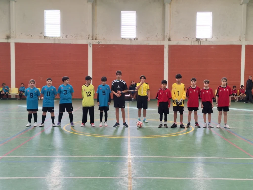 Duas equipas de futsal alinhadas lado a lado no centro do pavilhão, com árbitro e bola no círculo central, antes do jogo.