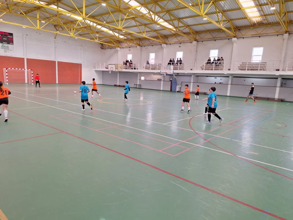 Jogo de futsal num pavilhão amplo, com equipas de camisolas azuis e laranja em campo e público sentado na bancada ao fundo.
