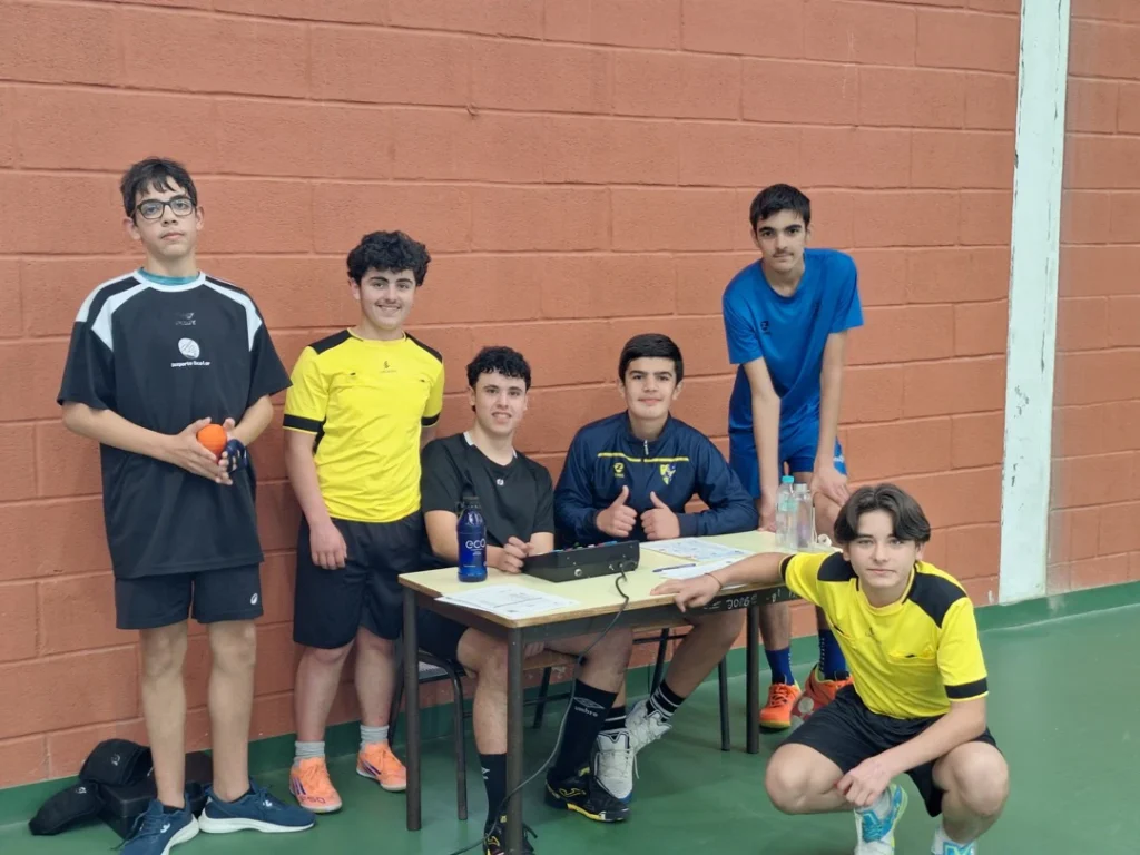 Seis alunos junto a uma mesa de arbitragem contra uma parede de tijolo, um deles com uma bola laranja na mão.