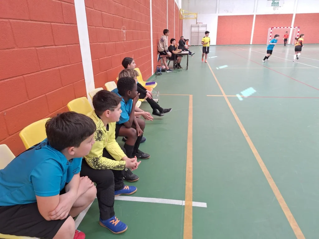 Alunos sentados no banco de suplentes, em cadeiras amarelas junto à parede do pavilhão, a ver o jogo de futsal.