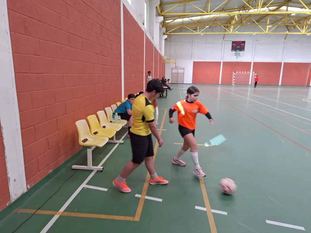 Dois alunos de futsal, um de camisola amarela e outro de camisola laranja, disputam a bola junto à linha lateral do pavilhão.