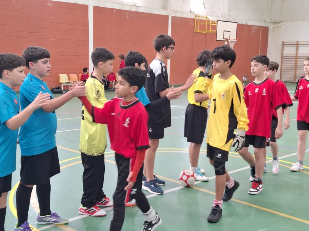 Alunos de equipas de futsal de camisolas vermelhas, azuis, amarelas e pretas cumprimentam-se antes do jogo, num pavilhão.
