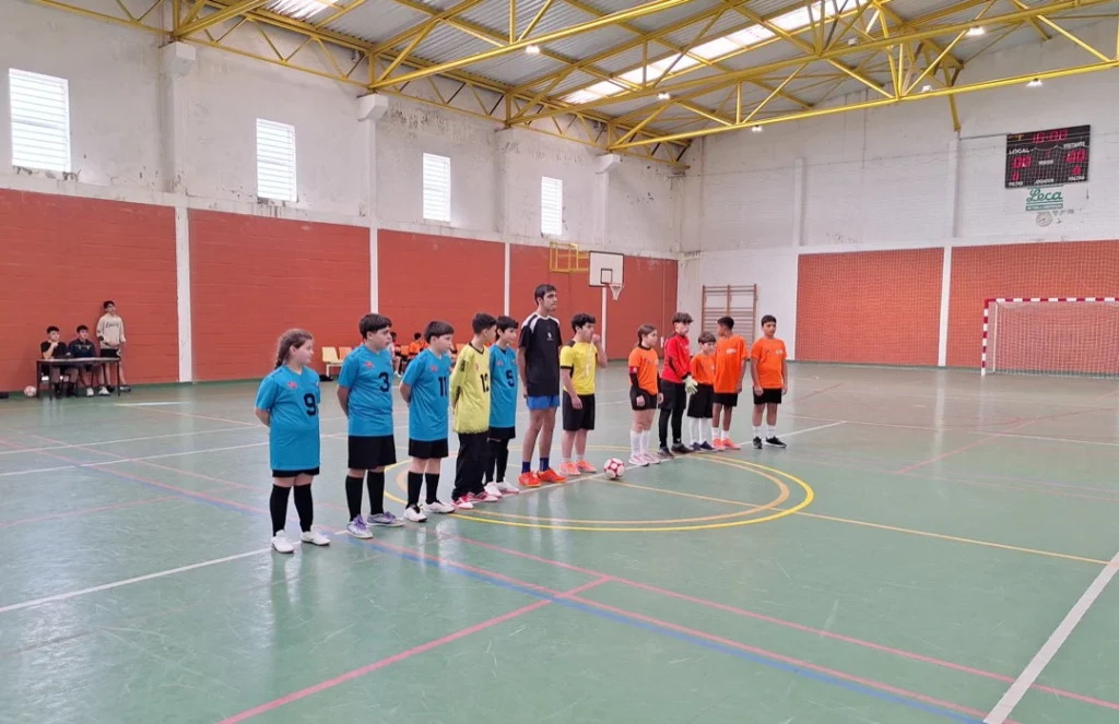 Duas equipas de futsal alinhadas no círculo central de um pavilhão, de camisolas azuis e laranja, antes do jogo.
