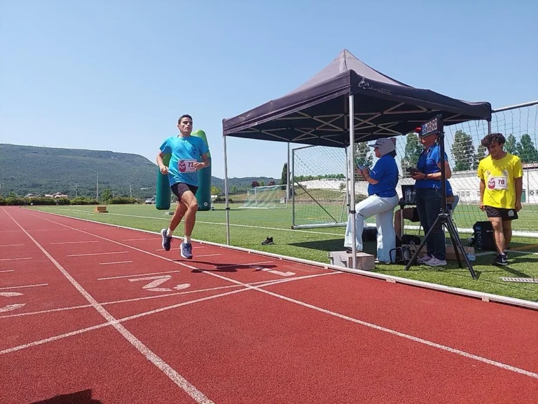 Atleta de dorsal 73 e camisola azul a correr numa pista vermelha; juízes sob uma tenda ao lado.