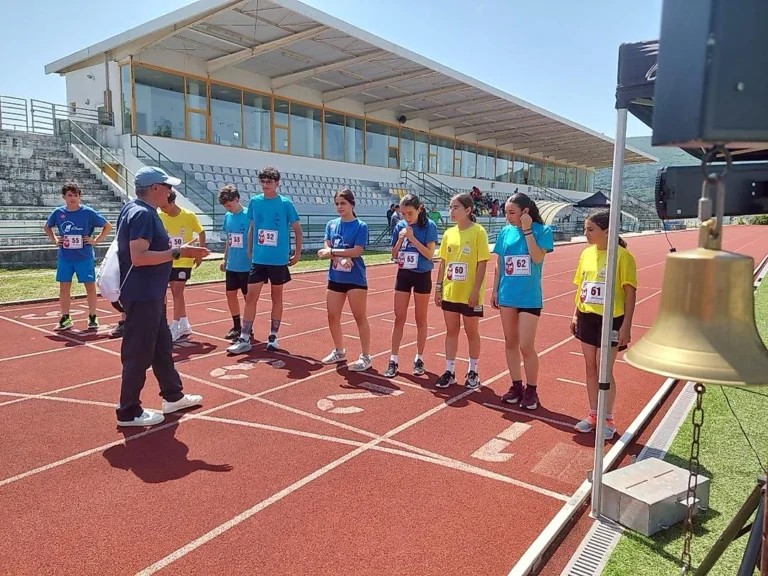 Adulto de boné dá instruções a alunos com dorsais numa pista de atletismo, junto a uma campainha.