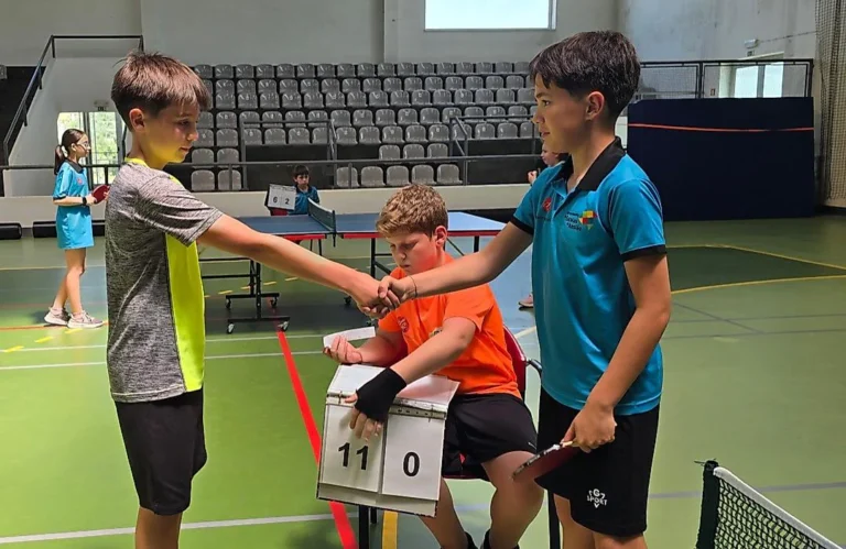 Dois jogadores apertam as mãos junto a um árbitro sentado, que mostra o marcador 11-0 num jogo de ténis de mesa.