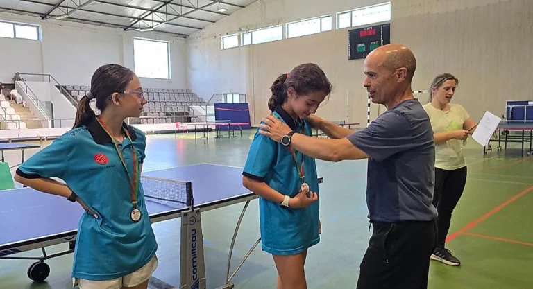 Adulto de t-shirt cinzenta a pôr a mão no ombro de uma aluna de polo azul com medalha, com outra aluna ao lado a observar.