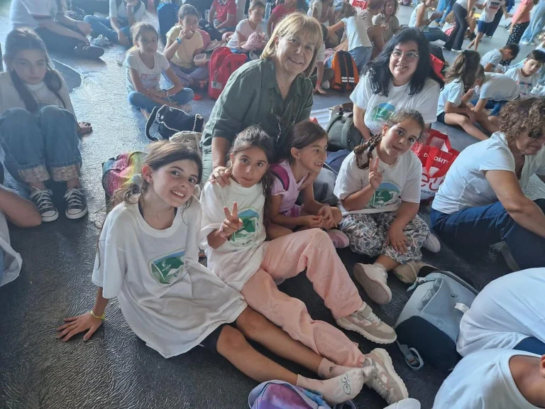 Alunos de t-shirt Eco-Escolas sentados no chão de um pavilhão; duas fazem o sinal de paz, com adultos atrás.