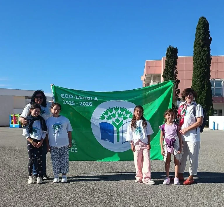 Grupo de alunos e adultos em frente à bandeira verde «Eco-Escola 2025-2026», com árvore e livro ao centro, num pátio ao sol.
