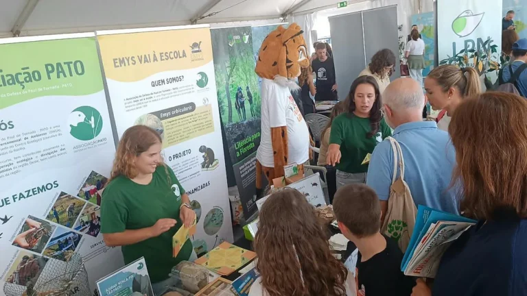 Stand da Associação PATO com painéis «Emys vai à escola»; ao lado, uma mascote laranja e castanho e visitantes.