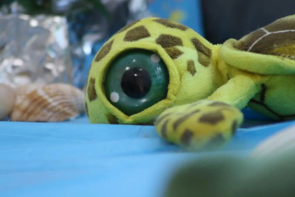 Peluche de tartaruga verde, de olhos grandes e manchas castanhas, deitado sobre uma superfície azul.