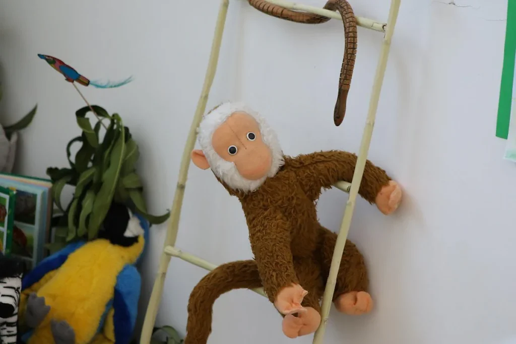 Peluche de macaco castanho, de cara clara e pelo branco à volta do rosto, a subir uma escada de canas.