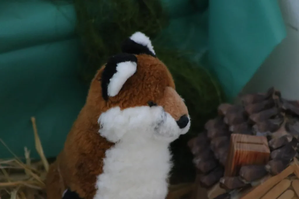 Peluche de raposa castanha e branca, de perfil, junto a pinhas e feno.