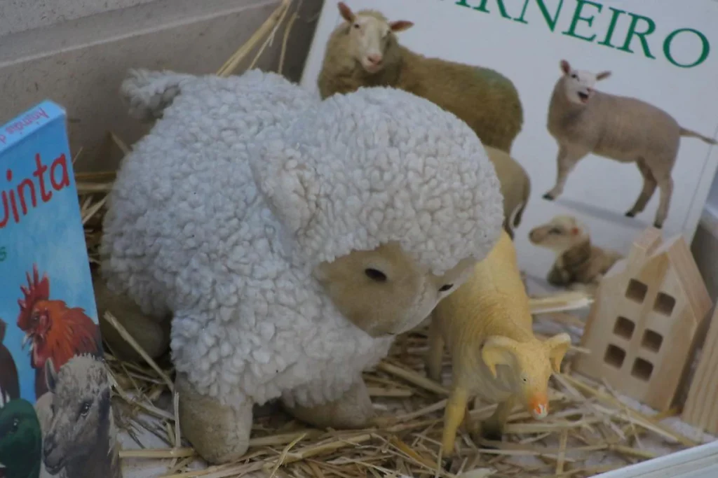 Peluche de ovelha branca e encaracolada, junto a um porco de plástico, com o livro «Carneiro» ao fundo.
